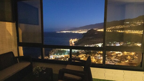 Una habitación con un ventanal grande con vistas a una ciudad costera iluminada de noche.