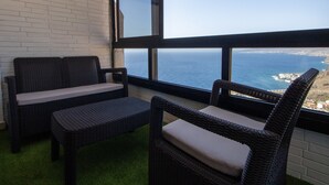 Una habitación con dos sillas de mimbre, una pequeña mesa, ventanas amplias con vista al océano y una pared de ladrillo blanco.