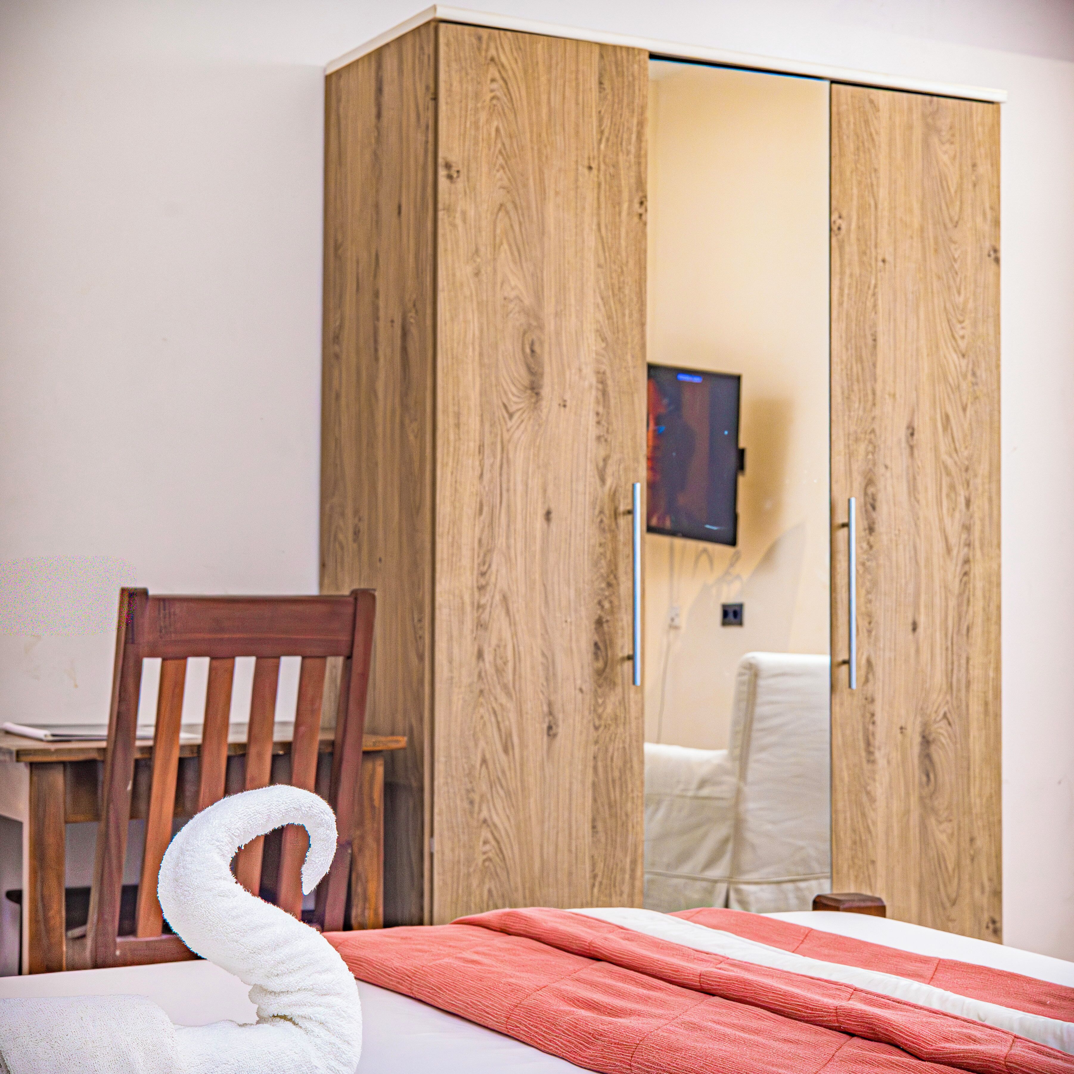 A bedroom with a wooden wardrobe, a chair, a bed with a red blanket, and a television on the wall.