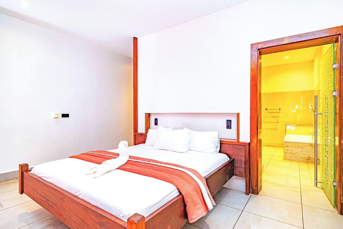 A modern hotel room with a wooden bed, white linens, and a glass door leading to a bathroom.