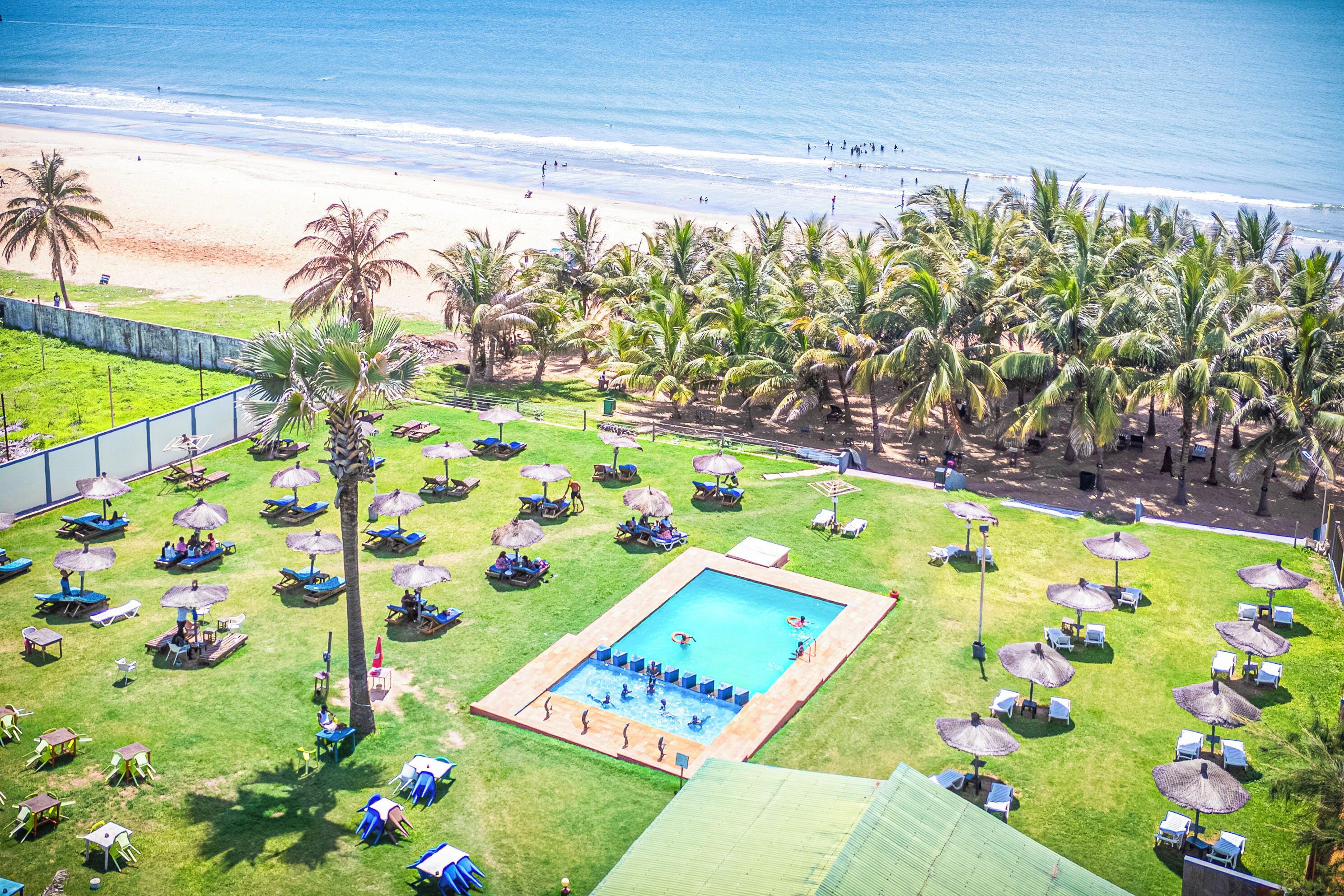 Zona de piscina con múltiples sillas de descanso y sombrillas, rodeada de palmeras y vista a la playa.