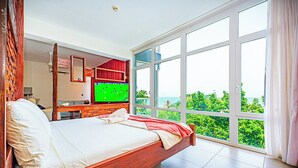 A bedroom with a large bed, a TV, and a view of greenery through large windows. - Kasumai Beach Resort (Serrekunda)