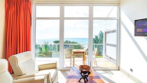 A room with a large sliding glass door leading to a balcony with a view of the ocean. There is a sofa and a small table with a decorative item on it. - Kasumai Beach Resort (Serrekunda)
