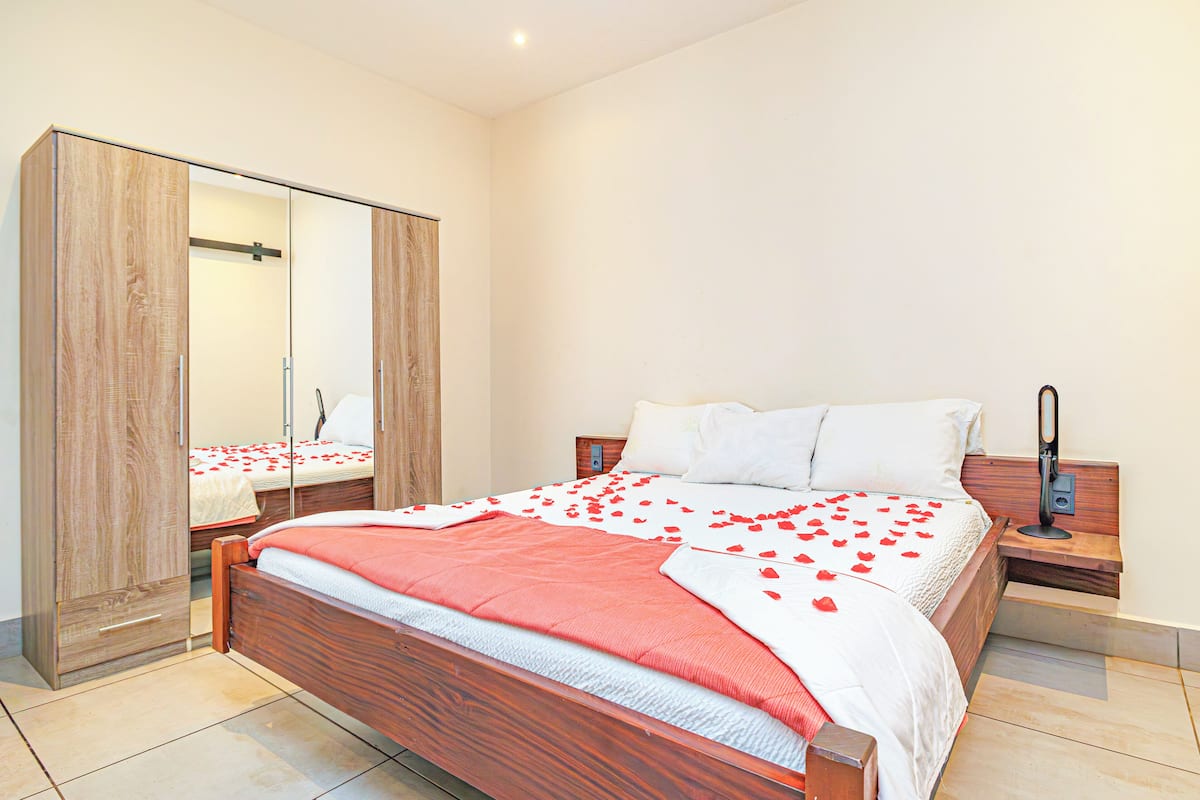 A neatly made bed with rose petals, a wooden wardrobe, and a bedside table with a lamp.