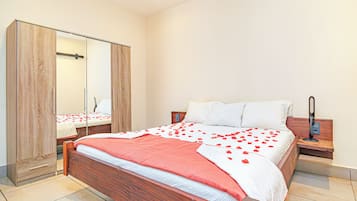 Una cama bien hecha con pétalos de rosa, un armario de madera y una mesita de noche con una lámpara.