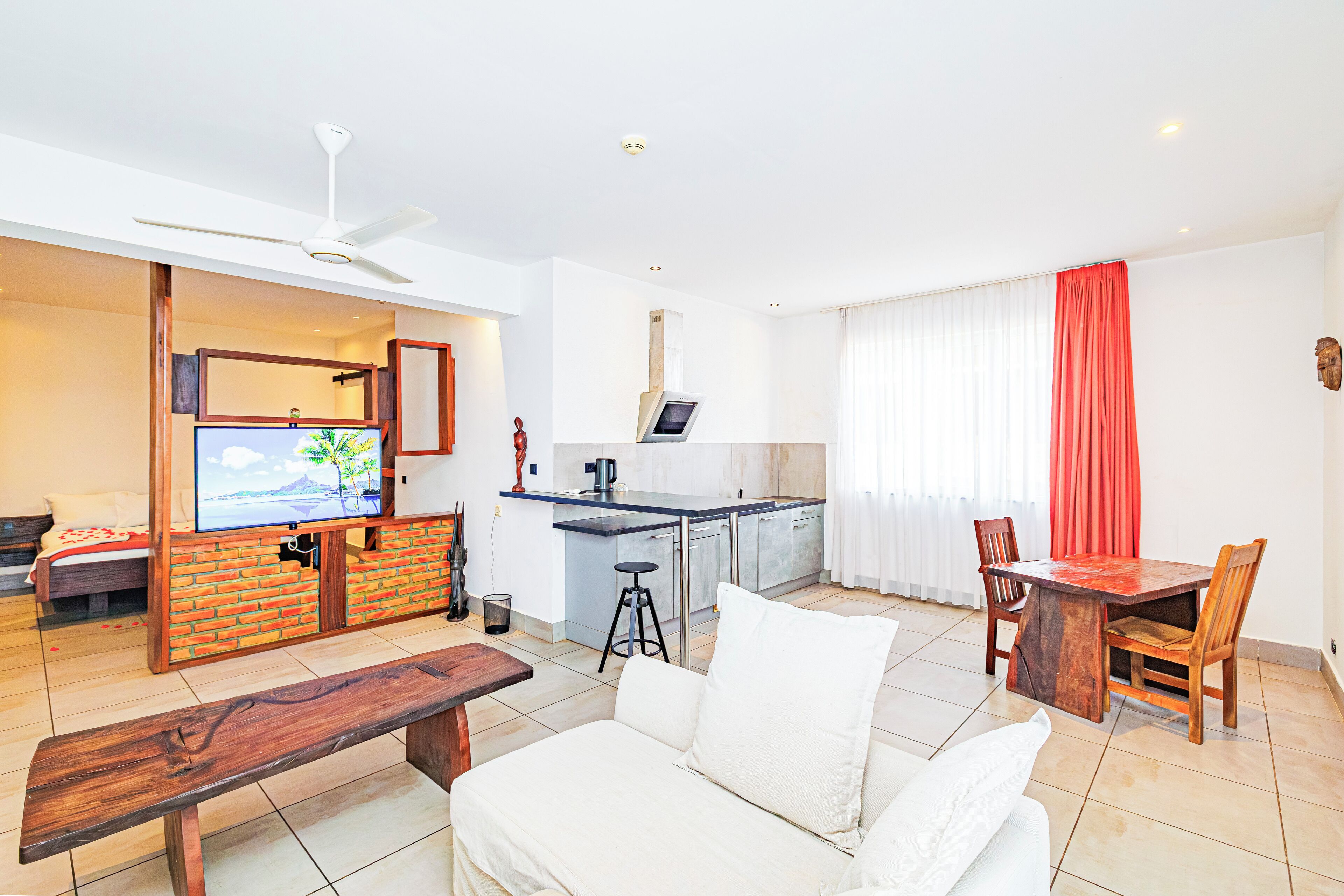 Una sala de estar moderna con un sofá, un banco de madera, una mesa de comedor con sillas, una zona de cocina con barra y un televisor montado en una pared de ladrillo.