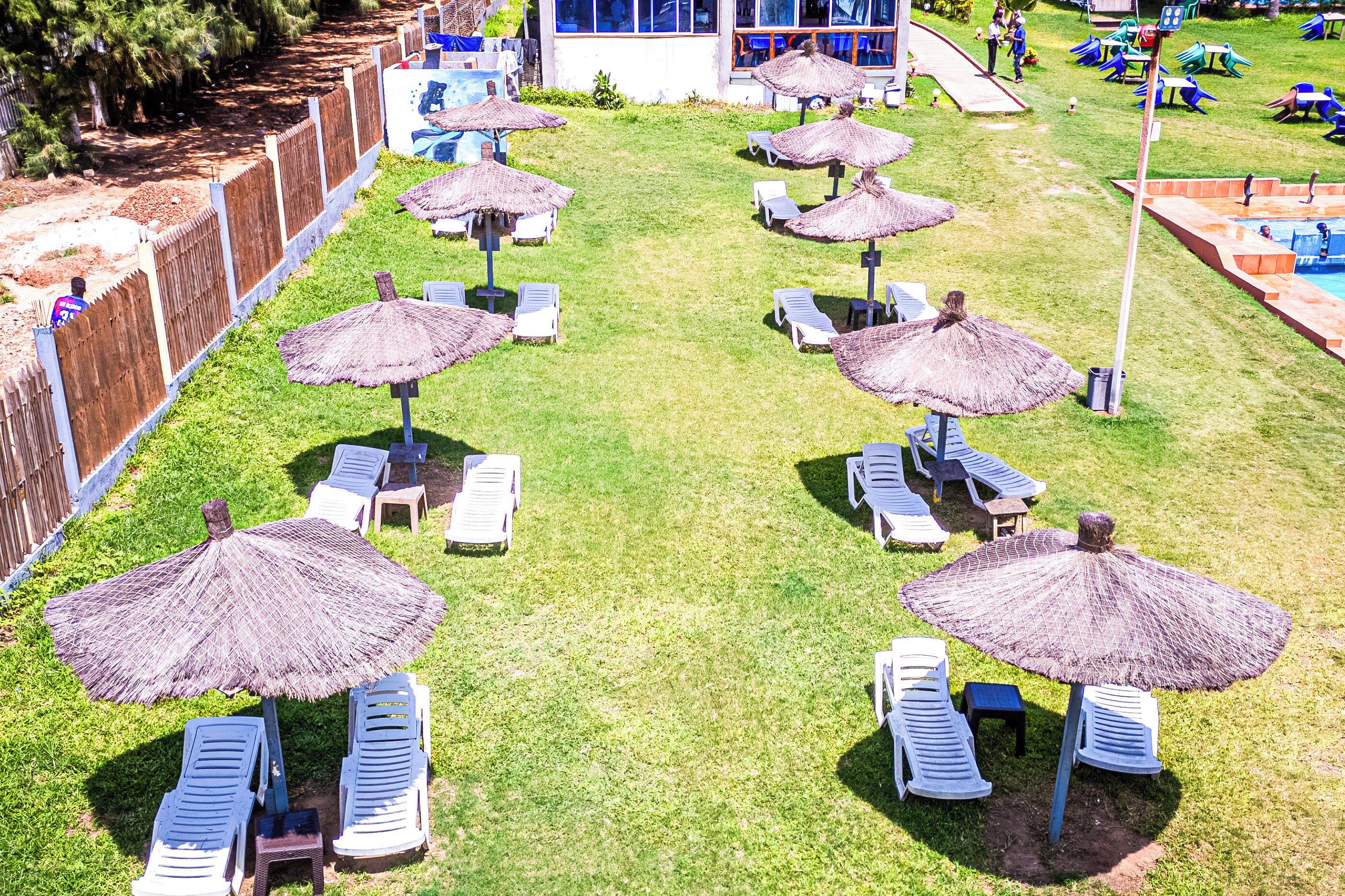 Un área junto a la piscina con sillas de descanso y sombrillas de paja, rodeada por una cerca de madera y un edificio con amplias ventanas.