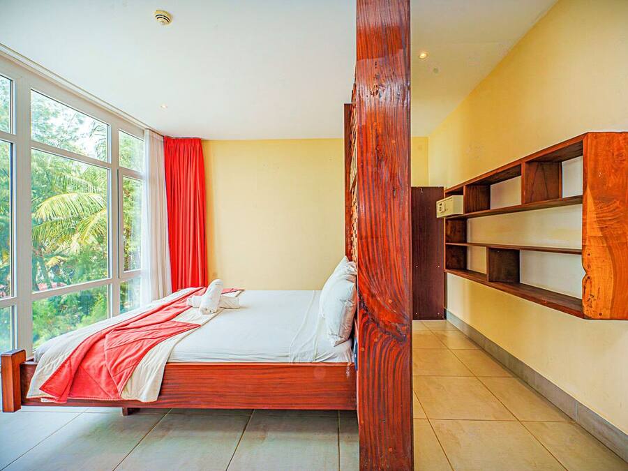 Un dormitorio con un ventanal amplio, una cama, una estantería de madera y una cortina roja.
