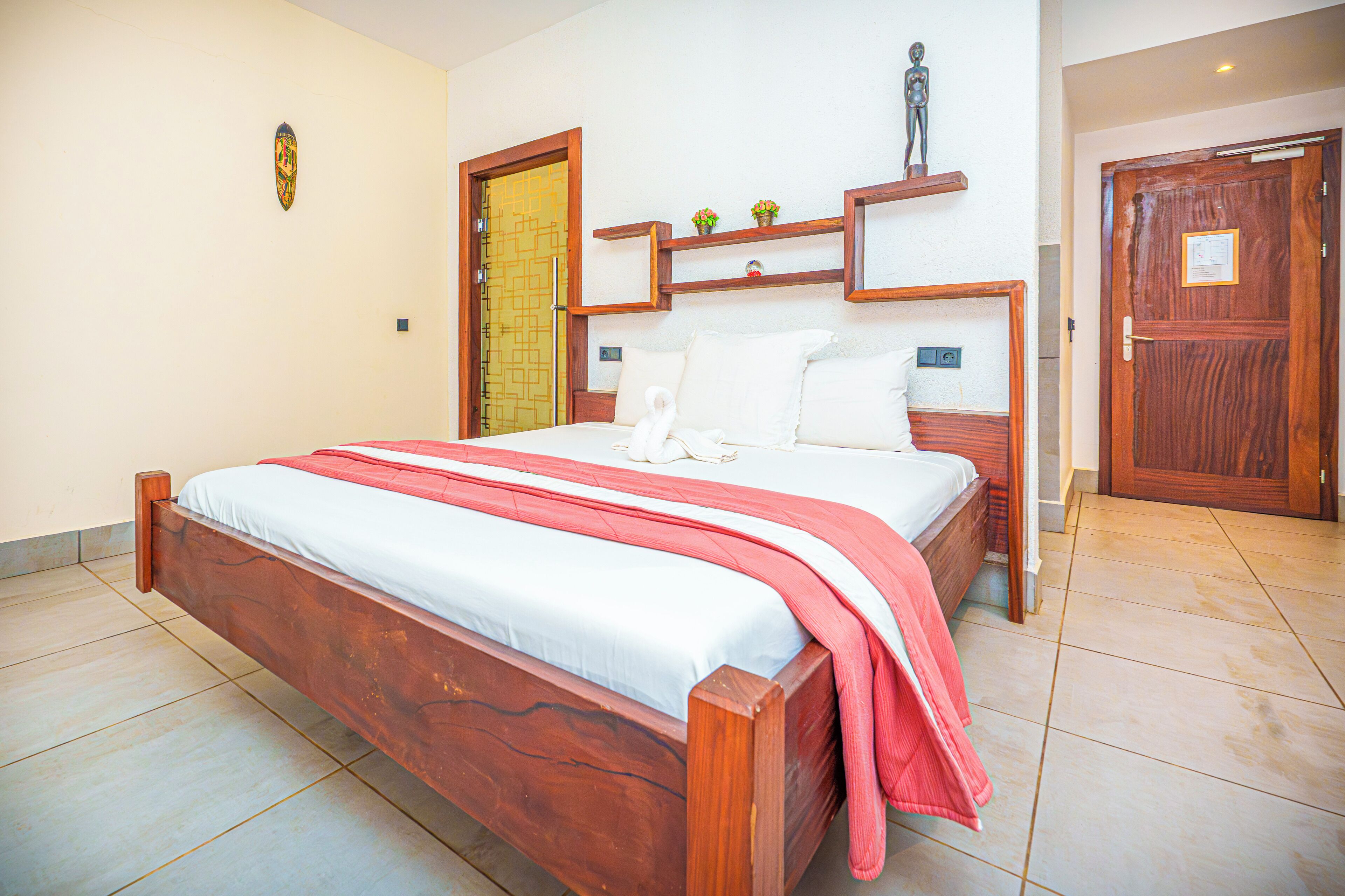 A bedroom with a wooden bed, white bedding, a red throw blanket, and a small shelf with decorative items.