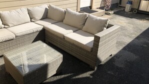 Un canapé d’angle en osier avec des coussins, un pouf assorti et une table basse avec un plateau en verre.