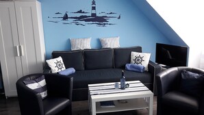 Una sala de estar con un sofá negro, dos sillones, una mesa de centro a rayas y un mural en la pared con un faro.