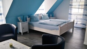 Un dormitorio con techo inclinado, una cama con ropa de cama azul, una mesita de noche, una silla y una mesa con un vaso.