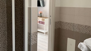 Un baño con ducha, inodoro y lavamanos. Hay un perchero con toallas y una mesita con un florero.