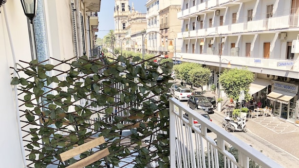 Un balcón con planta en maceta, banco de madera y vista a un paisaje urbano que incluye una torre y edificios.