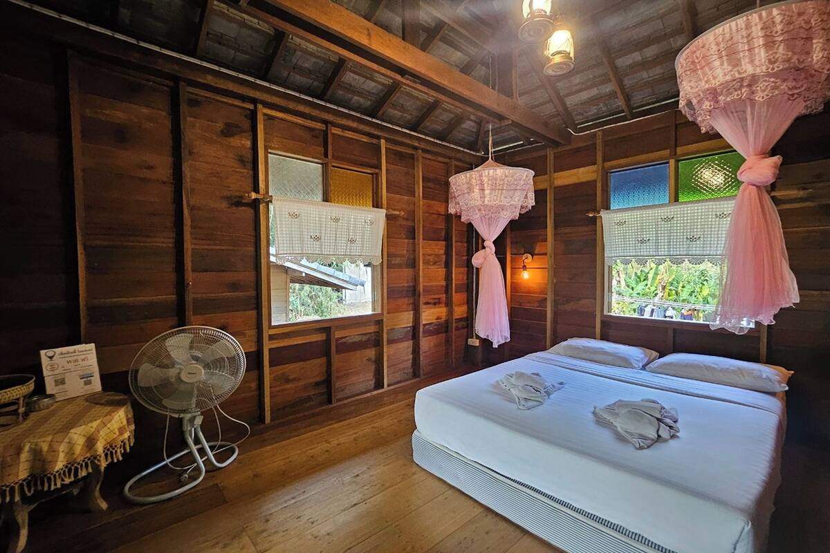 Ein Zimmer in einer Holzhütte mit einem Bett, einem Ventilator und einem Fenster mit Blick auf Grünflächen.