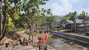 A wooden deck leading to a stone bridge over a stream, surrounded by greenery and a small building with a green roof.