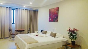 A neatly made bed with a heart-shaped pillow arrangement, a small table with a chair, a potted plant, and a painting on the wall. - Village Plaza Hotel (Barbacena)