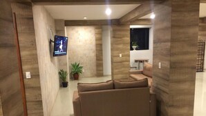A modern living room with a beige sofa, a flat-screen TV mounted on the wall, and a potted plant. - Village Plaza Hotel (Barbacena)