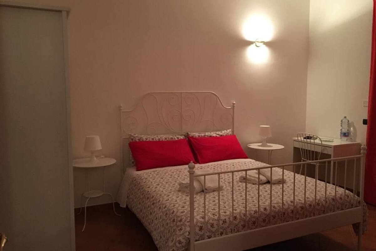 A bedroom with a white metal bed frame, a red and white patterned bedspread, two red pillows, a bedside table with a lamp, and a wall-mounted light fixture.