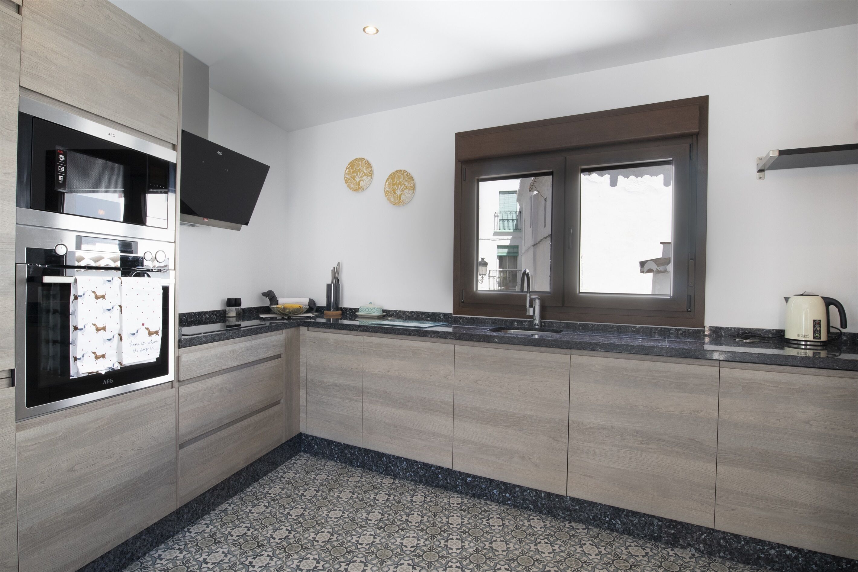 Een moderne keuken met een inbouwoven, een magnetron en een spoelbak. Er is een raam met uitzicht naar buiten, en een decoratief bord aan de muur.