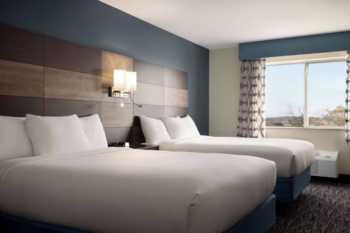 A hotel room with two beds, a wall-mounted light fixture, a window with curtains, and a view of the outdoors.
