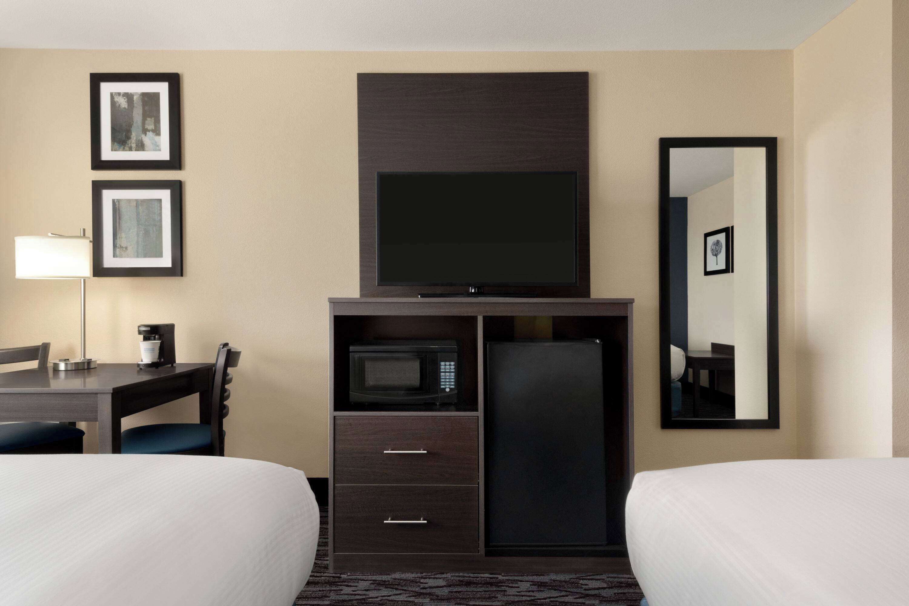 A hotel room with two beds, a desk with a lamp, a microwave, a refrigerator, and a flat-screen TV mounted above the desk.