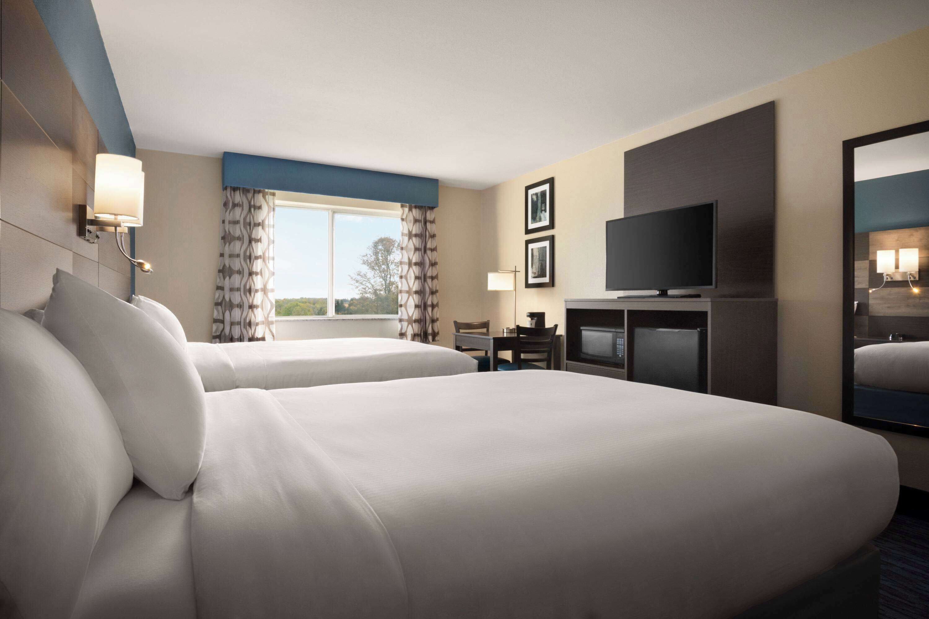 A hotel room with two beds, a television, a desk, and a large window with curtains.