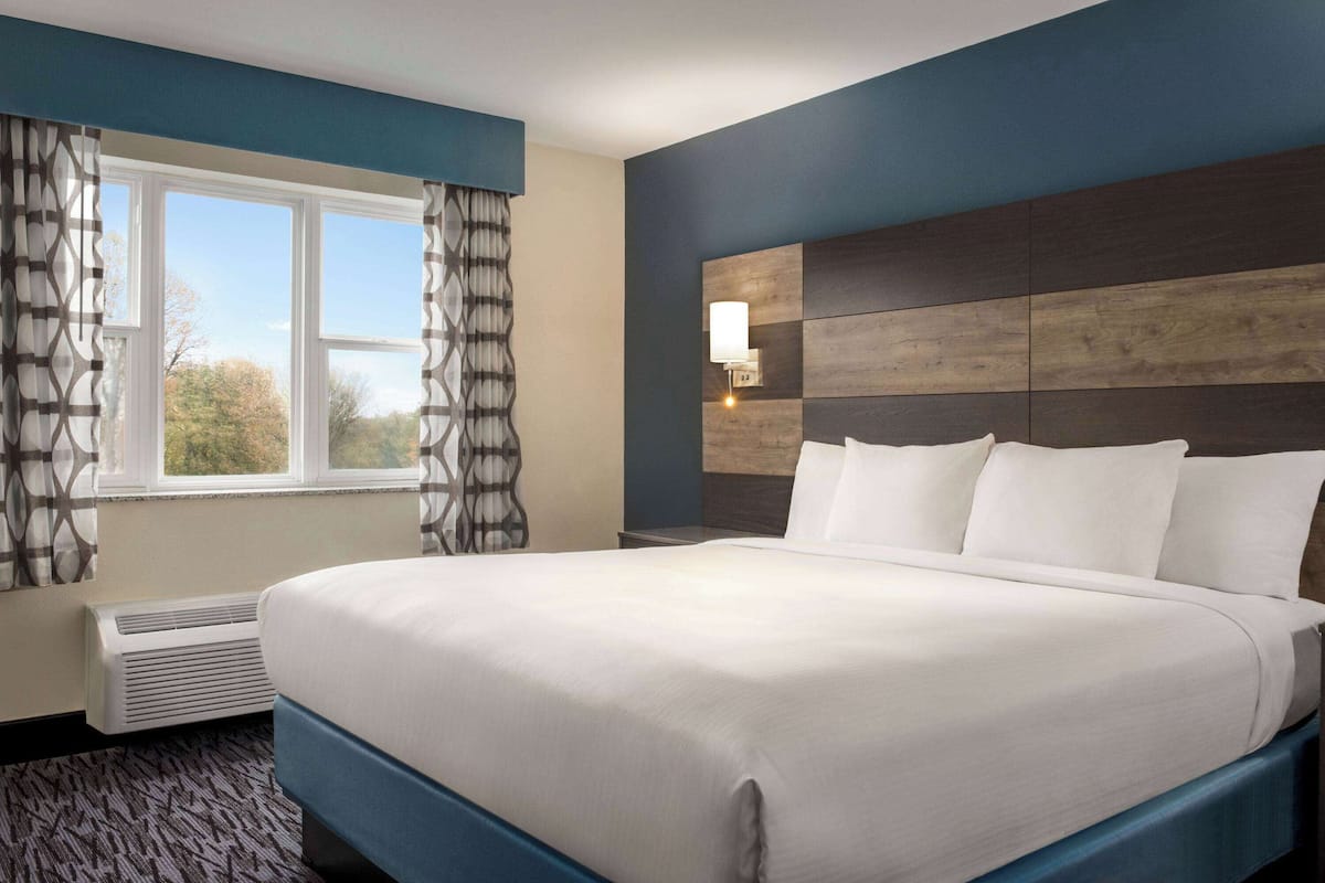 A hotel room with a large bed, a window with curtains, a wall-mounted lamp, and a built-in heater.