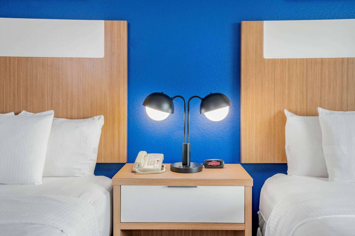 A hotel room with two beds, a bedside table with a phone, a lamp, and a small table with a red object.