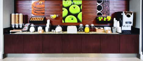 Zona de recepción de hotel con un mostrador que exhibe diferentes alimentos y bebidas, un cartel grande con el texto "LA QUINTA" y una máquina de café.