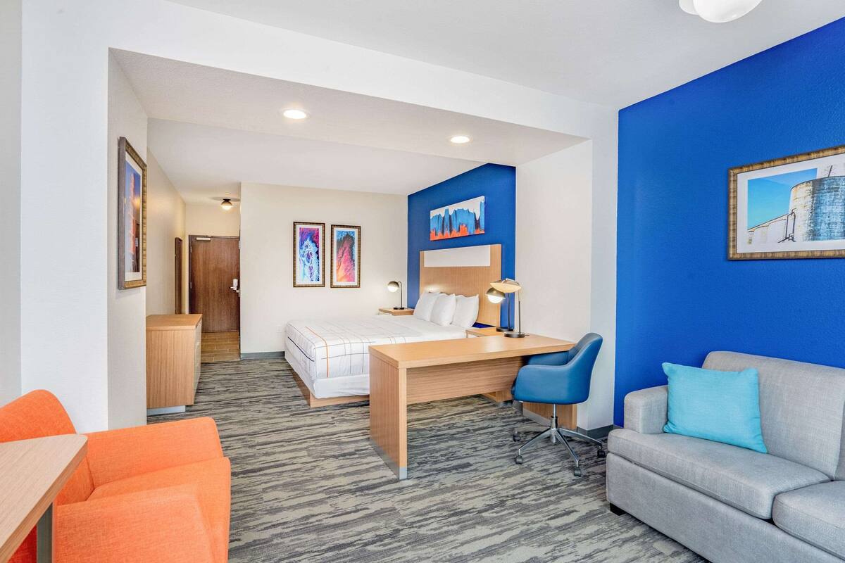 A hotel room with a bed, desk, chair, and a couch. There are framed pictures on the wall and a door leading to another room.