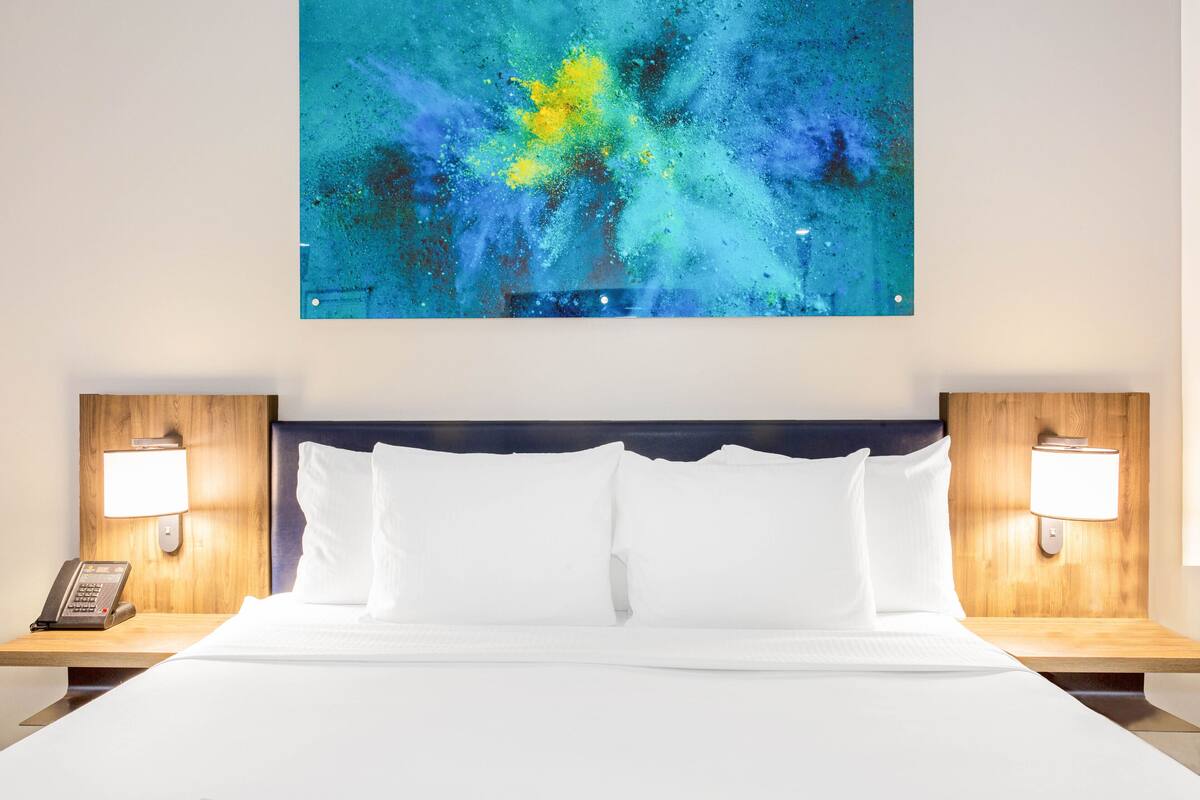 A hotel room with a large bed, white linens, two bedside tables with lamps, and a wall-mounted abstract painting.