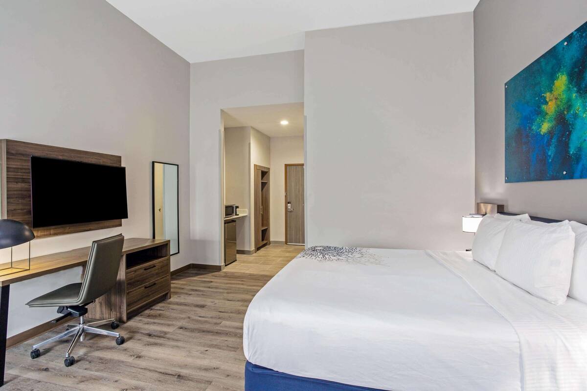 A modern hotel room with a large bed, a desk with a flat-screen TV, a chair, and a painting on the wall.