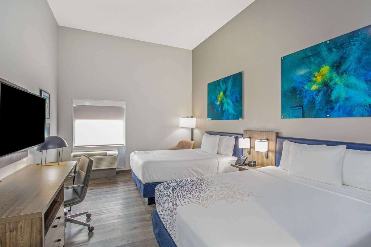 A hotel room with two beds, a desk, a chair, a TV, and artwork on the walls.