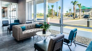 Un moderno vestíbulo de hotel con un sofá color beige, sillas azules, una pequeña mesa redonda con una planta y ventanas amplias que ofrecen vistas a palmeras y un estacionamiento.