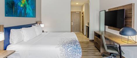 A modern hotel room with a large bed, a desk with a computer, a flat-screen TV, and a colorful abstract painting on the wall.
