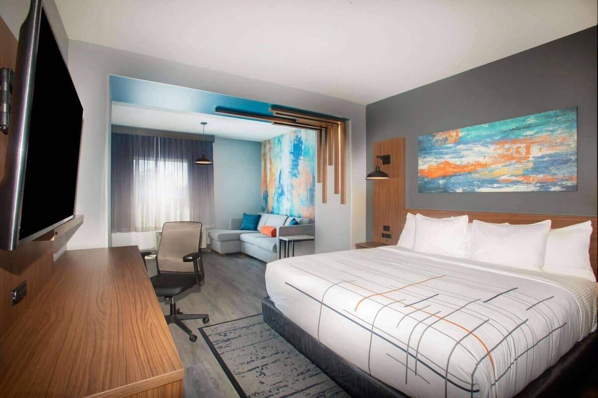 Une chambre d’hôtel avec un grand lit, un bureau, une chaise, une télévision, un tableau et une fenêtre avec des rideaux.