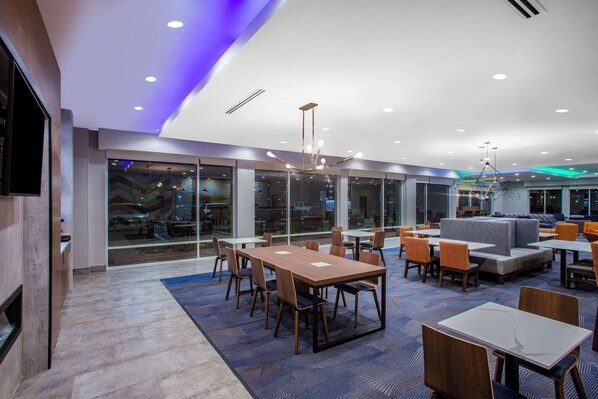 Un comedor moderno con mesas y sillas, ventanas amplias y una alfombra azul.
