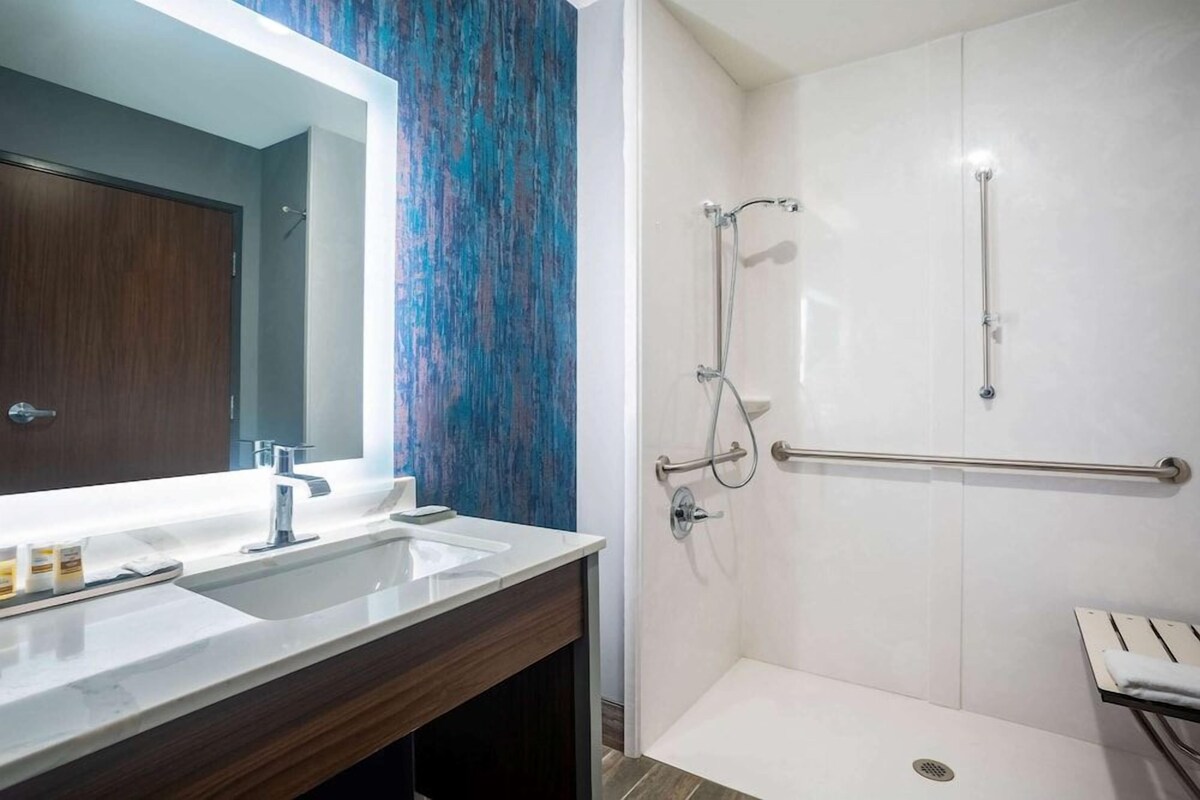 Un baño con un espejo grande, dos lavamanos, ducha con barras de apoyo y un perchero para toallas.
