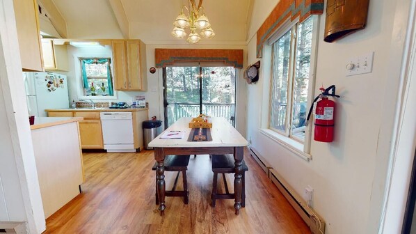 Une cuisine de taille réduite avec des meubles en bois, un plan de travail blanc, une table à manger en bois avec deux chaises, une fenêtre donnant sur des arbres et un extincteur fixé au mur.