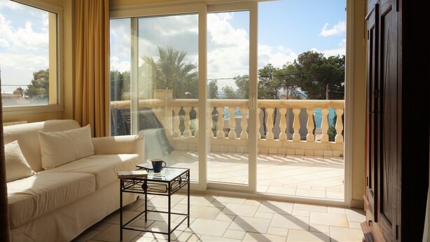 Una stanza con un divano beige, un tavolino con una tazza e vista su un balcone con ringhiera e alberi.