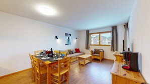 A cozy living room with a wooden dining table set for four, a sofa, a coffee table, and a TV on a wooden stand.