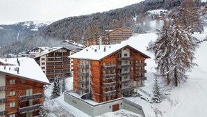 A snow-covered mountain lodge with multiple wooden buildings, balconies, and a ski slope in the background.