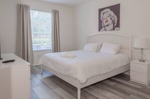 Une chambre à coucher blanche, avec un lit, une table de chevet avec une lampe, une fenêtre avec des rideaux et un tableau accroché au mur.