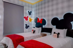 Quarto de hotel com duas camas, cada uma com almofada do Mickey Mouse, manta vermelha e uma janela grande.