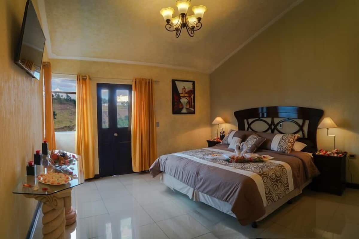 A bedroom with a bed, bedside lamps, a TV mounted on the wall, a door with curtains, and a painting on the wall.
