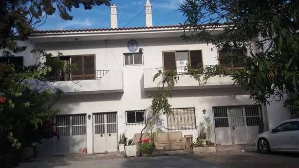 A two-story building with a white facade, multiple windows, and a red-tiled roof. There is a car parked in front and a tree to the right. - Hostel Pereira is on the first floor of a detached villa (Loulé)