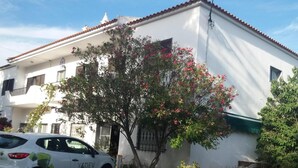 A white building with a red-tiled roof, a parked white car, and a tree with pink flowers. - Hostel Pereira is on the first floor of a detached villa (Loulé)