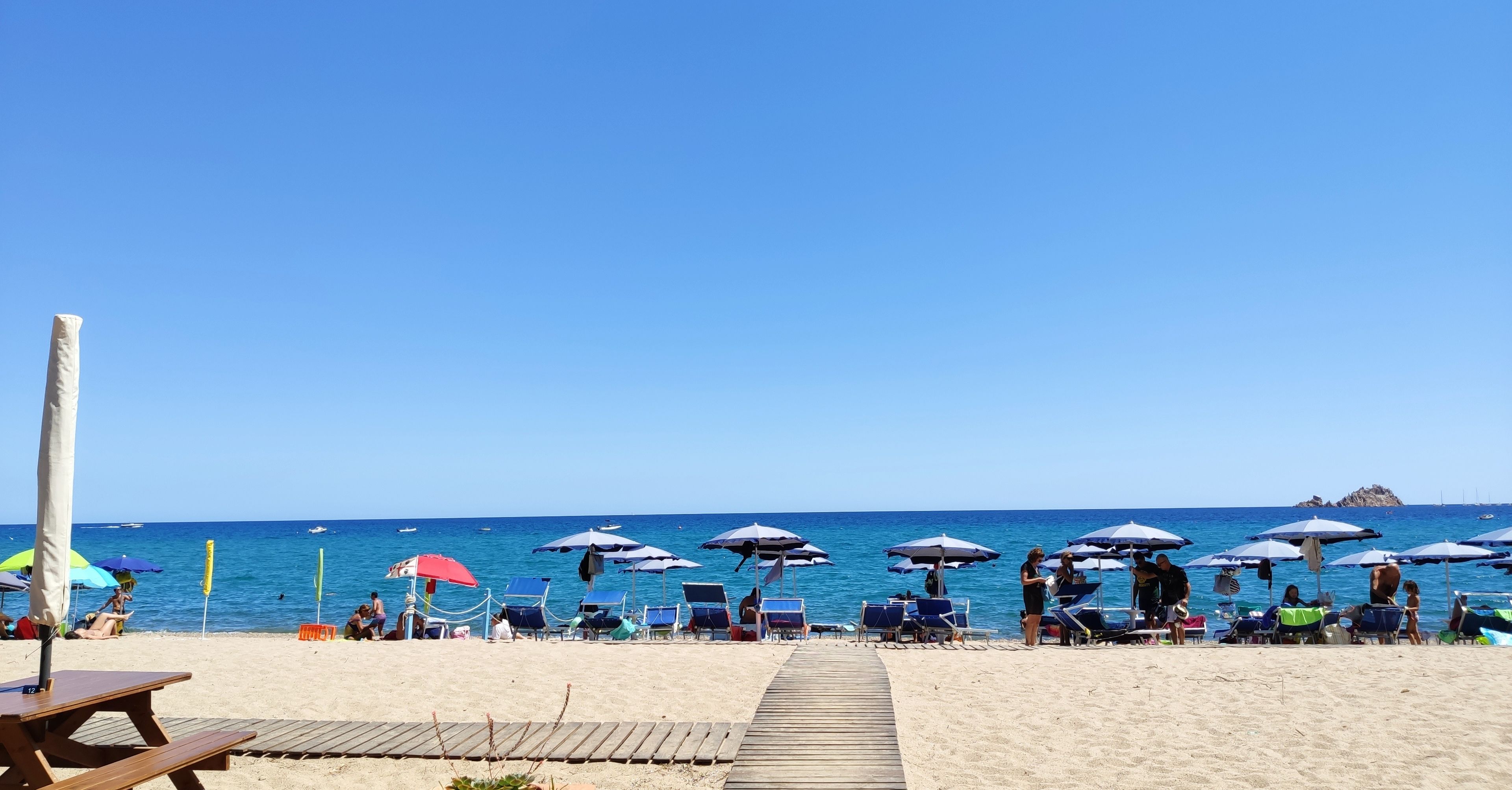 A beach with rows of sun umbrellas and lounge chairs, a wooden walkway leading to the water, and a clear view of the sea.
