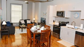 A modern kitchen with stainless steel appliances, a dining area with wooden furniture, and a living room with a sofa and a TV.